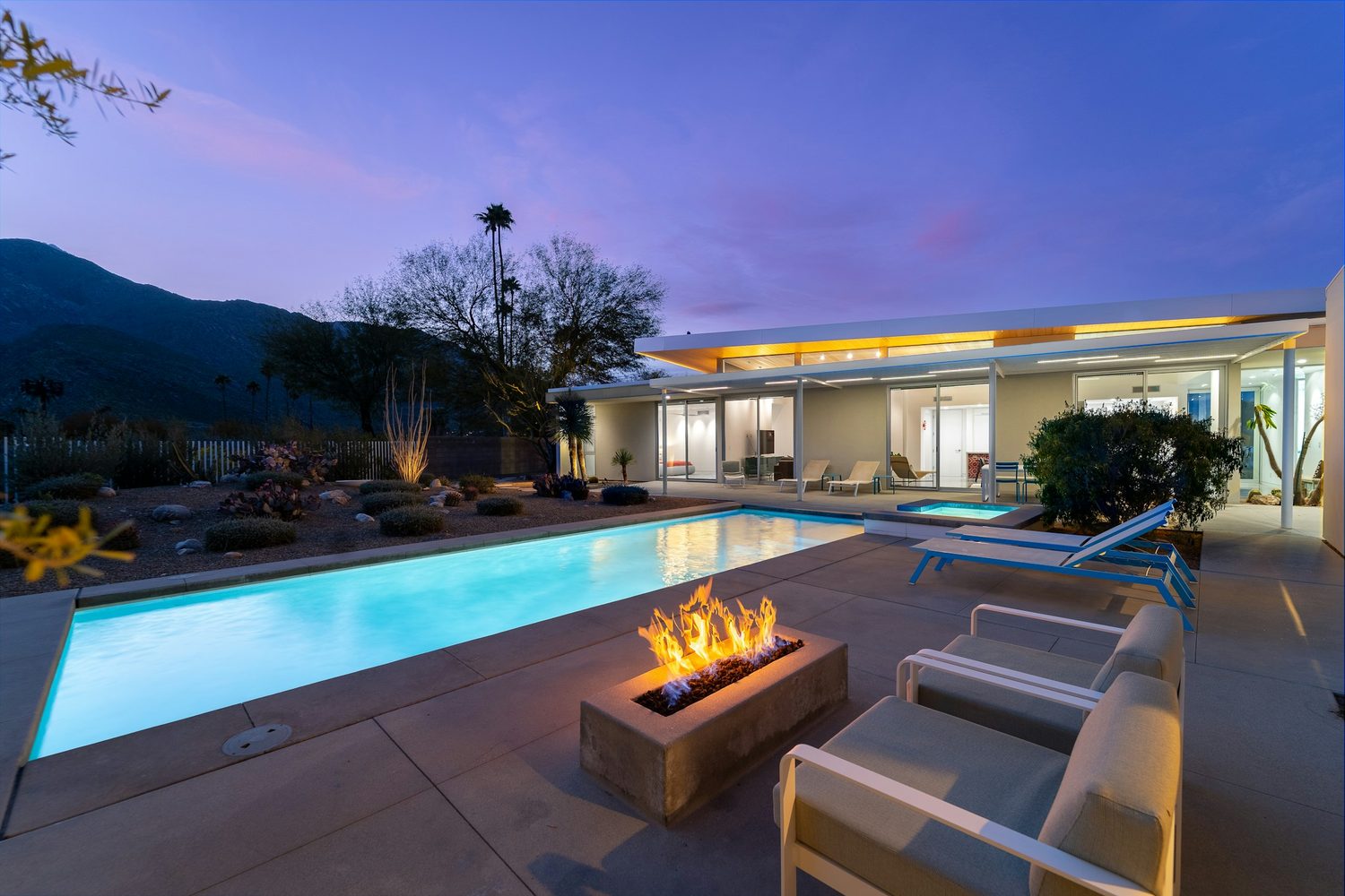 A desert home at dusk, pool and fire feature lit, mountain silhouette behind